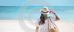 Happy traveler woman in white dress and hat enjoy beautiful sea view, young woman standing on sand and looking ocean at tropical