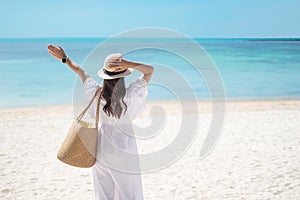 Happy traveler woman in white dress and hat enjoy beautiful sea view, young woman standing on sand and looking ocean at tropical