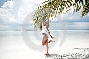 Happy traveller woman. Blonde Bikini Model posing by palm on sand on tropical beach vacation. Sexy slim girl in white swimwear.