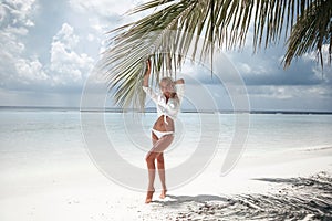 Happy traveller woman. Blonde Bikini Model posing by palm on sand on tropical beach vacation. Sexy slim girl in white swimwear.