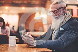 Happy trendy man drinking coffee and using smartphone in bar cafeteria indoor - Fashion senior guy having fun with web app -