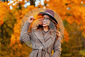 Happy trendy young woman in glasses in a vintage hat in an elegant coat poses with an orange-yellow autumn leaf outdoors in a park