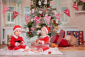Happy twin babies dressed as Santa Claus are sitting near Christmas tree at home in living room