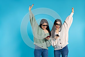 Happy twin girls in 3D glasses with smartphones on a blue background. Hands up and empty side space