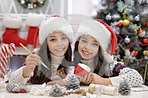 Happy twin girls in Santa hats preparing for Christmas sittind at the table, looking at the camera