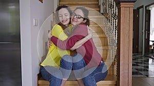 Happy twin sisters smiling hugging looking at camera sitting on stairs at home indoors. Positive beautiful slim