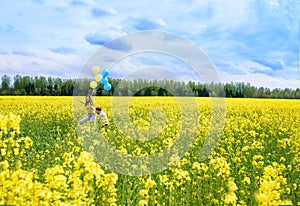 happy two children run on a yellow field, blooming rapeseed. blue sky and clouds. Blue and yellow balloons, concept of