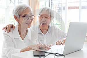 Happy two senior asian woman,sisters or friends talking and enjoying using laptop computer together at home,smiling elderly people