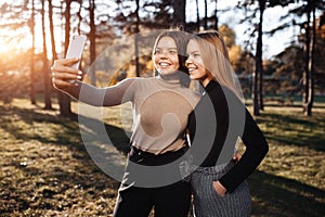 Happy two young twin sisters in casual outfit with bright smile take selfie with smartphone at the park