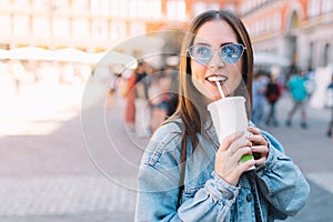 Happy urban woman with blue sunglasses enjoying her morning drinking a soda in styrofoam cup with straw. Pretty girl in the street