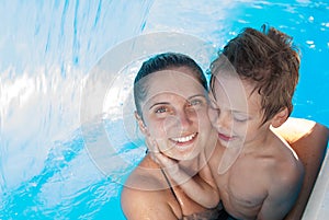 Happy vacation of young family mother and child embracing in azure swimming pool with jet waterfall during summer vacation leisure
