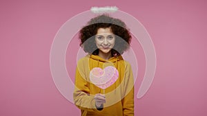 Happy Valentine`s Day! Angelic girl afro hairstyle with nimbus holding pink heart, looking playful flirting and sending air kiss,