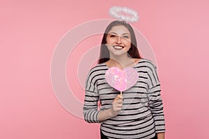 Happy Valentine`s day! Angelic woman with holy nimbus holding pink paper heart and smiling to camera