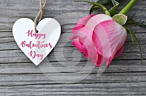 Happy Valentine`s Day greeting card with pink rose and decorative white heart with text on old wooden background.14th of February.