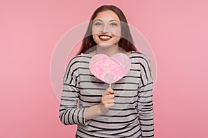 Happy Valentine`s day! Joyful pretty woman holding pink paper heart and looking at camera with toothy smile