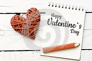 Happy Valentine`s Day message. Text on spiral notepad`Happy Valentine`s Day` with pen beside it on the right and woven hearts on t