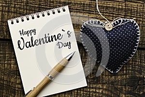 Happy Valentine`s Day message. Text on spiral notepad`Happy Valentine`s Day` with pen beside it on the right and woven hearts on t