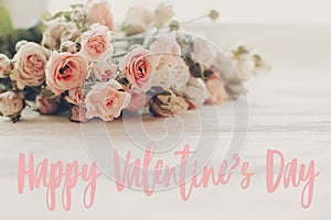 Happy Valentine`s Day text sign on pink small roses on wooden background in light. Tender Flowers image. Valentines day floral