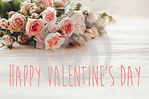 Happy Valentine`s Day text sign on pink small roses on wooden background in light. Tender Flowers image. Valentines day floral