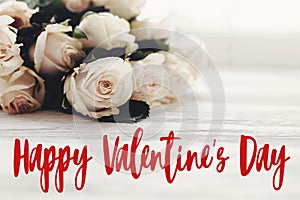 Happy Valentine`s Day text sign and white roses bouquet in light on wooden background. Valentines day floral greeting card