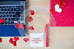 Happy Valentine`s day text written on white stickers, pc, red pen, gift box decorated by red hearts on wooden table in office.