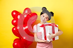 Happy Valentines day. Cute asian girl hugging surprise gift from boyfriend, standing near lovely red heart balloons and