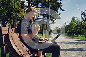 happy victorious bearded man sit on the bench outdoor while look at laptop gesturing yes win winner