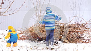 Happy village children in bright and warm clothes play around campfire from branches against gray fence in garden. Children are ha