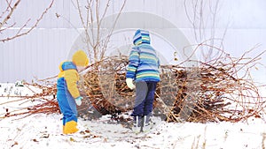 Happy village children in bright and warm clothes play around campfire from branches against gray fence in garden. Children are ha
