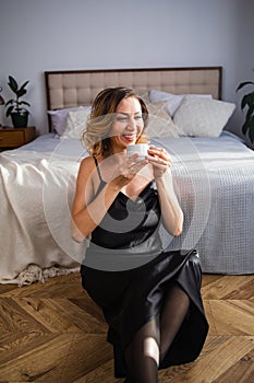 Happy middle-aged blond woman holding a cup of tea or coffee looking at the camera in cozy home