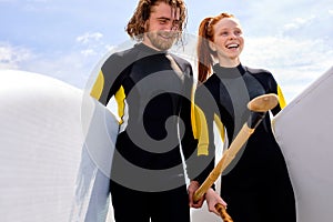 Happy watersport couple with SUP paddle boards going to sea, in swimwear wetsuit