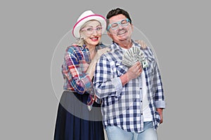 Happy wealthy family, adult man and woman in casual checkered shirt standing pickaback together, holding fan of dollar, toothy