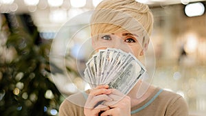 Happy wealthy successful middle aged adult caucasian business woman looking at camera holding fan of money hiding behind