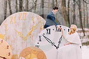 Happy wedding couple softly holding hands at the big vintage clocks in autumn forest. Creative decorations