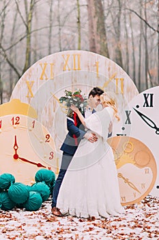 Happy wedding couple softly hugs at the big vintage clocks in autumn forest. Creative decorations