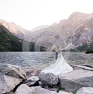 Happy wedding couple is standing in front of lake surrounded with mountains on the huge rock