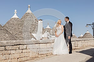 Happy wedding couple standing on street near conical roofs of trullo, meeting eyes. Young bride hugging with groom.
