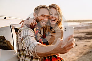 Happy white couple taking selfie photo by car while walking at seashore