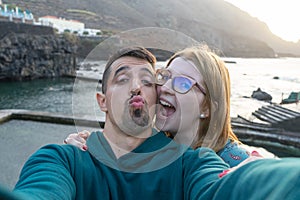 Happy wife with glasses laughing, giving hug with tenderness her latin husband, sending kiss to camera, flirting in coast. Friends