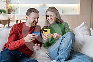 Happy wife and husband spending money in online shops using smartphone with credit card at home