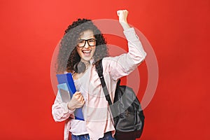 Happy winner celebrating victory. Young curly student woman wearing backpack glasses holding books and tablet over isolated red