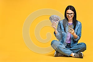 Happy winner! Portrait of a smiling cheerful woman using smartphone isolated over yellow background. Sitting on the floor in lotus