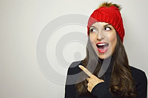 Happy winter woman with red hat looking to the side and pointing with her finger your product on gray background. Copy space.