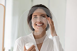 Happy woman in bathrobe holds jar applying cream on cheek