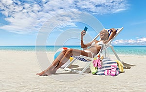 Happy woman at the beach on beach deck chair, sunbathing, uses mobile phone, in a sunny day with blue sky, concept a summer beach