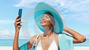 Happy woman at the beach side wearing bikini, blue sun hat and pareo and using mobile phone in a sunny day with blue sky.Concept