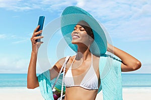 Happy woman at the beach side wearing bikini, blue sun hat and pareo and using mobile phone in a sunny day with blue sky.Concept photo