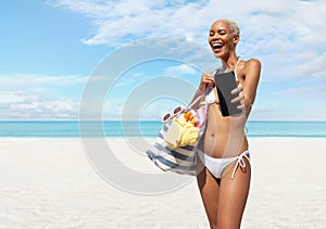 Happy woman at the beach side wearing bikini holding a beach bag and showing mobile phone in a sunny day with blue sky. Concept of