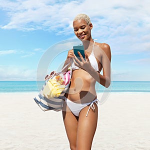 Happy woman at the beach side wearing bikini holding a beach bag and using mobile phone in a sunny day with blue sky. Concept of