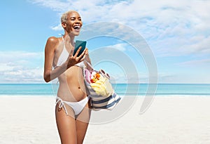 Happy woman at the beach side wearing bikini holding a beach bag and using mobile phone in a sunny day with blue sky. Concept of